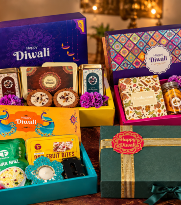 Four festive corporate Diwali gift hampers featuring colorful gift boxes, traditional sweets, dry fruit bites, snacks, and elegant green packaging, arranged closely together with glowing diyas and marigold flowers.